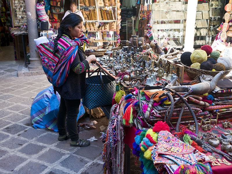 A Peruvian woman in Cusco is shopping for goods while carrying a baby on her back